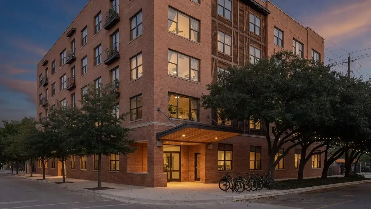 Street-corner view of The Alder at dusk with a timber-slat entry canopy, glowing windows and live oaks
