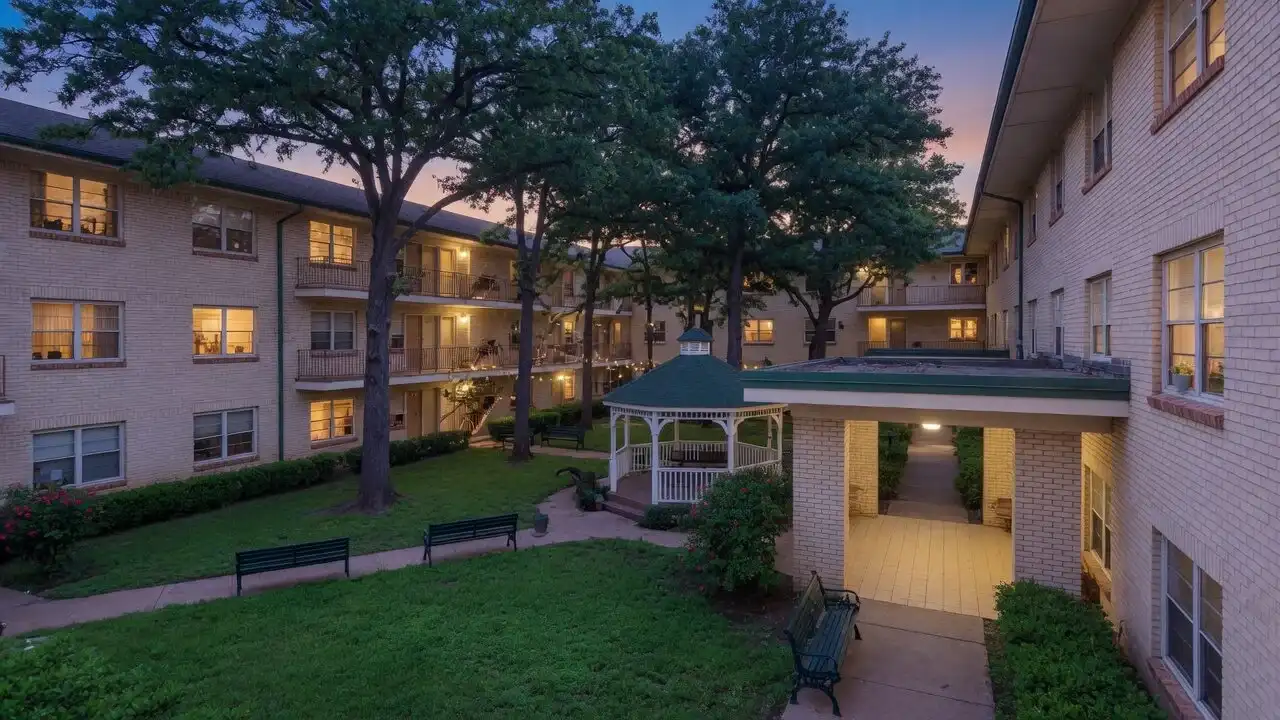 Cream brick garden-style buildings framing a green courtyard with a gazebo and shade trees at dusk